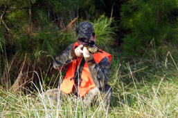 A man in a camouflage outfit with an orange vest is leaning in the grass, pointing his firearm towards the camera. 